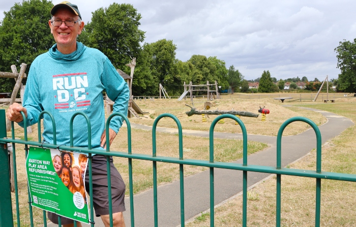 Cllr Chris Lloyd standing by the green gated entrance to the Barton Way Play Area. A poster with a QR code advertising the consultation is in the foreground. Play equipment is in the background.