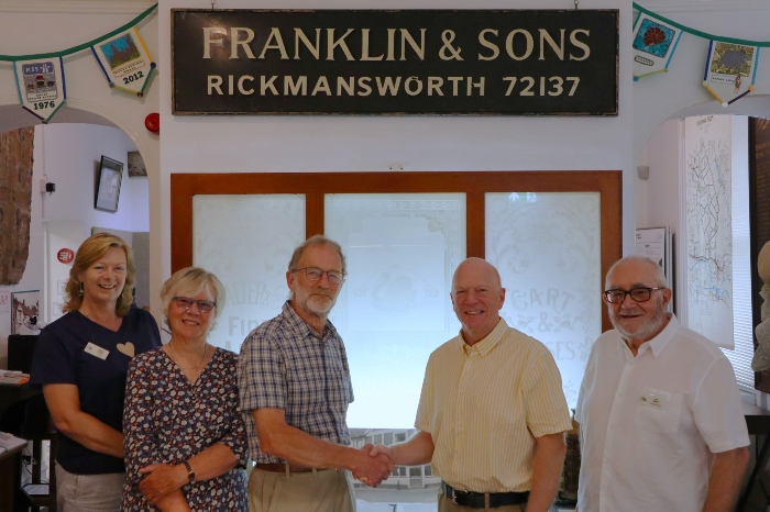 The Museum's chairman, Fabian Hiscock, shaking hands with Council Leader, Cllr Stephen Giles-Medhurst, surrounded by museum volunteers underneath an old sign that reads 'Franklin & Son Rickmansworth 72137'.