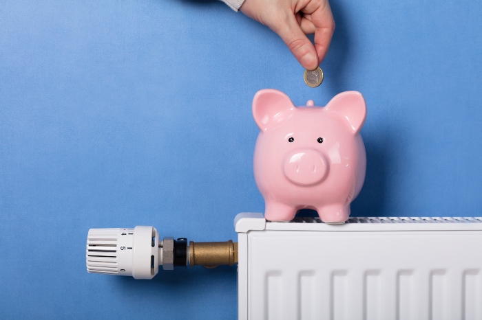 A hand putting a pound coin in a piggy bank that's sitting on a radiator. The wall is painted blue.