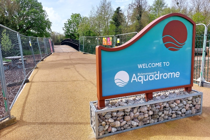 Rickmansworth Aquadrome welcome sign in the foreground with the red bridge in the background. Heras fencing to the side.
