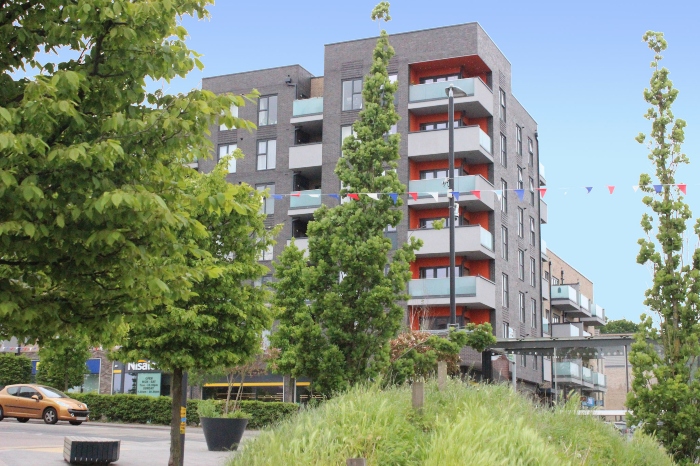 An apartment block with shops on the ground floor in the background with trees and an orange car driving on the road in foreground.