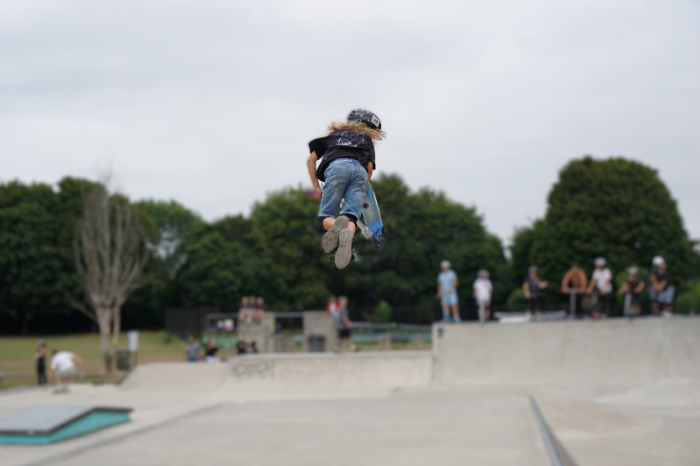 A young person, wearing a helmet, in the air above a skate ramp, with a scooter out to one side