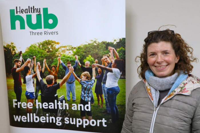 Image of a woman stood smiling next to a pull up banner that reads 'Healthy Hub Three Rivers Free health and wellbeing support'.